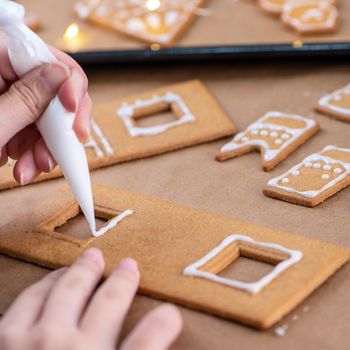 Young woman is decorating Christmas Gingerbread House cookies biscuit at home with frosting topping in icing bag, close up, lifestyle.