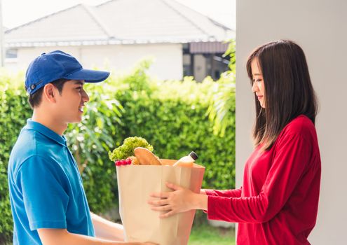 Asian young delivery man in uniform making grocery fast service giving fresh food in paper bag to woman customer receiving at house door under pandemic coronavirus, Back to new normal concept