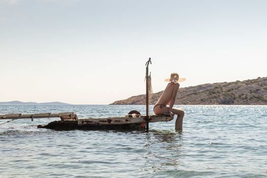 View of unrecognizable woman wearing big summer sun hat tanning topless and relaxing on old wooden pier in remote calm cove of Adriatic sea, Croatia.