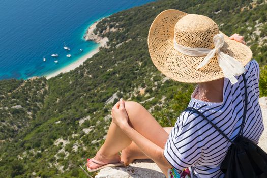 Active sporty woman on summer vacations sitting on old stone wall at Lubenice village, wearing straw hat and beach backpack enjoying beautiful coastal view of Cres island, Croatia.