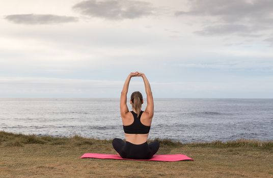 Woman in black sportswear exercising outdoors on a pink mat with the sea in the background.