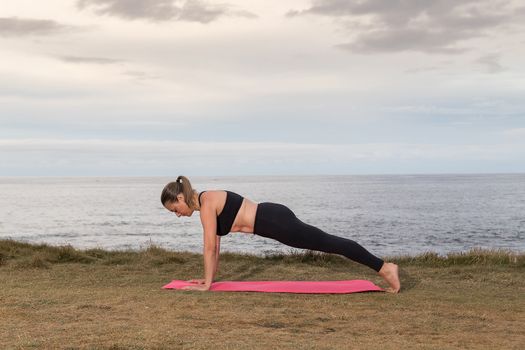 Woman in black sportswear exercising outdoors on a pink mat with the sea in the background.