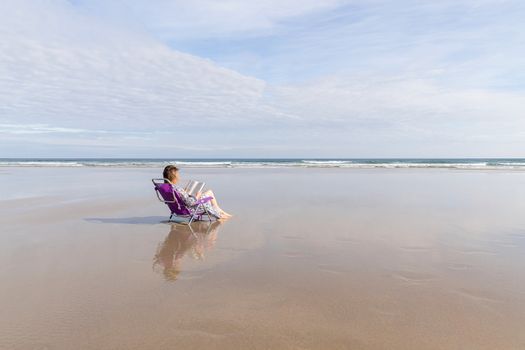 Barefoot woman reading a book sitting on a chair on the beach. Copy space. Social distance concept.