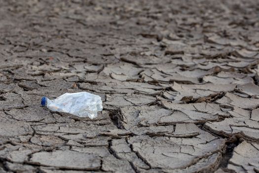 empty crumpled plastic bottle on dry cracked mud desert like gray background, close-up with selective focus and blur