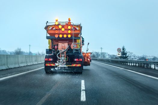 View from the car behind orange highway maintenance truck spreading de-icing salt and sand, crystals dropping on the ice covered asphalt road.