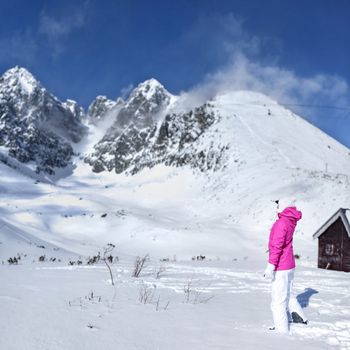 Young woman in pink ski jacket, gloves, hat looking at the top of snow covered Lomnicky stit peak, shading her eyes with hand on a sunny day.  Skalnate sedlo ski resort, Slovakia.