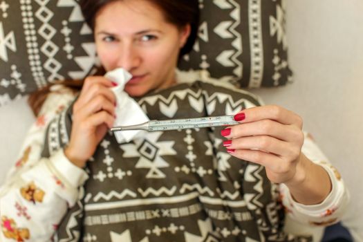 Sick woman lying in the bed, covered with duvet, holding paper tissue handkerchief in one hand and in other mercury thermometer showing temperature over 38 degrees Celsius indicating high fever. 