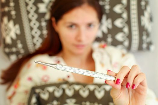 Sick young woman lying in the bed, covered with duvet,  holding mercury thermometer showing temperature over 38 degrees Celsius indicating high fever. 