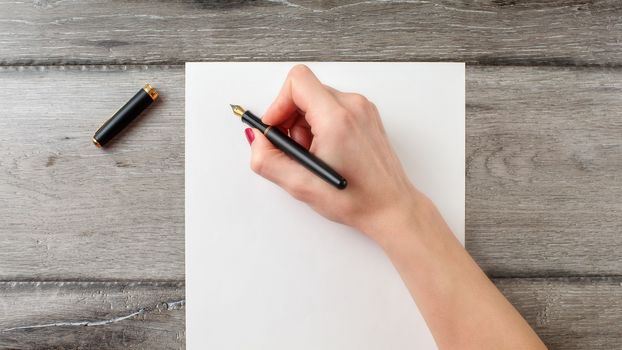 Woman's hand holding black fountain pen ready to write on blank paper on gray wooden desk