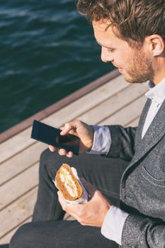Professional man on lunch break from work using cellphone eating sandwich outside by the canal water. Caucasian young adult holding smart phone mobile app.