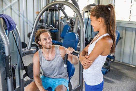 Fitness gym trainer talking to man training on workout equipment machine indoors. Couple happy working out together.