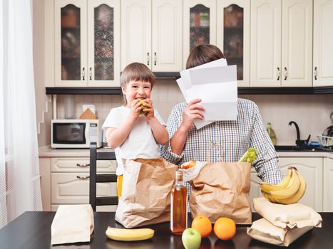 Family sorts out purchases in the kitchen. Father and son shows expensiveness of products in shopping bags. Grocery delivery in conditions of quarantine because of coronavirus COVID19.