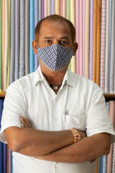Portrait of shopkeeper looking at camera in cloth store wearing protective medical mask to prevent coronavirus or covid-19 and standing with arms crossed.
