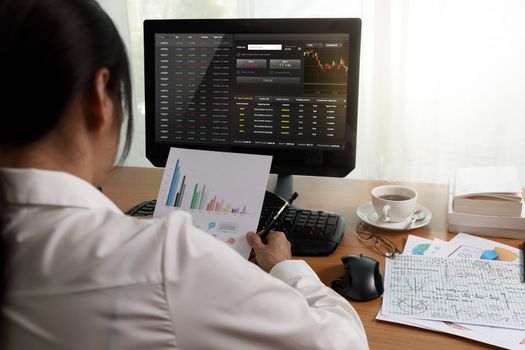Rear view of businesswoman working in office with computer holding chart report paper and looking. businesswoman analysis project statistics data on PC screen and paper. Business and finance concept.