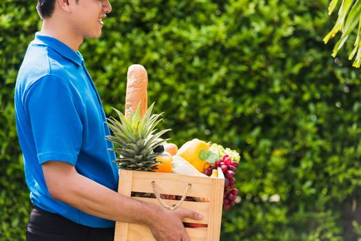 Asian man farmer wears delivery uniform he holding full fresh vegetables and fruits in crate wood box in hands ready give to customer harvest organic food on the garden place green leaves background