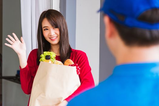 Asian young delivery man in blue uniform making grocery service giving fresh vegetables and fruits and food in paper bag to woman customer at front house after pandemic coronavirus, Back to new normal