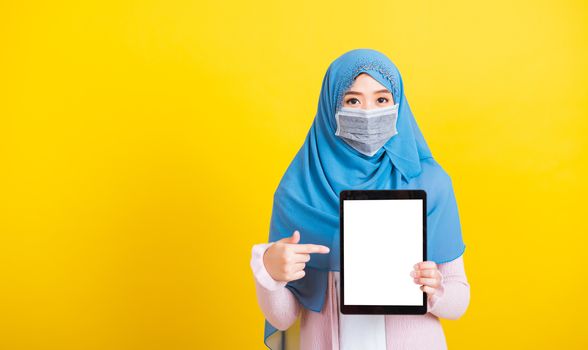 Asian Muslim Arab, Portrait of happy beautiful young woman Islam religious wear veil hijab and face mask protect quarantines disease coronavirus show blank screen tablet isolated yellow background