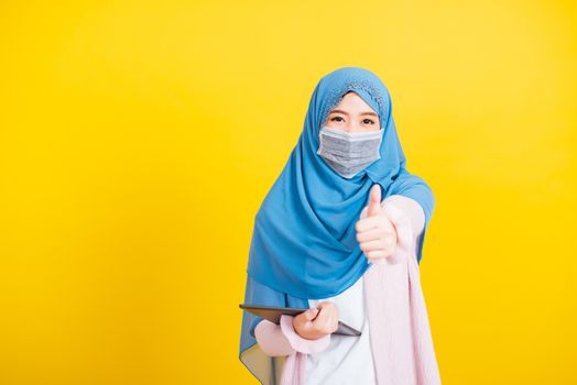 Asian Muslim Arab, Portrait of happy beautiful young woman Islam religious wear veil hijab and face mask protect she quarantines disease coronavirus show finger thumb up for good sign isolated yellow