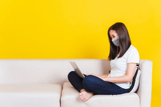 Portrait Asian of beautiful young woman sitting on sofa wearing medical face mask protective she work from home with laptop computer during Coronavirus studio shot isolated on yellow background