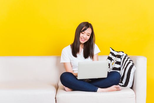 Portrait Asian of happy beautiful young woman work from home she sitting on sofa using laptop computer in house living room studio shot isolated on yellow background