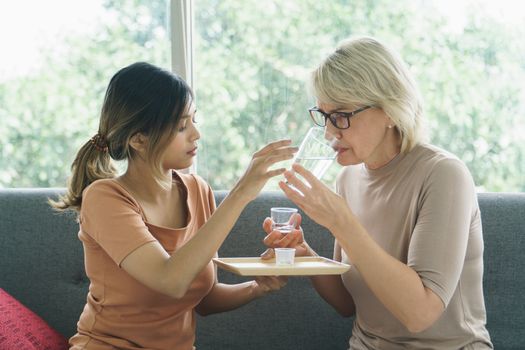 Asian Growing daughter giving medical pill with  glass of water, sick older Caucasian mother.concept family mix skin, love, care, time-sharing, adoption, stepmother, daughter-in-law
