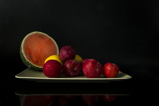 still life with a green bowl on a reflective background with lemons, plums and half a water meleon