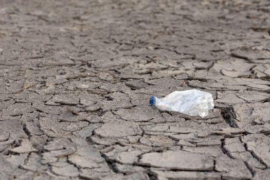 empty crumpled plastic bottle on dry cracked mud desert like gray background, close-up with selective focus and blur