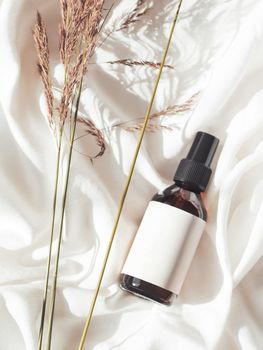 Concept of apothecary cosmetic. Herbs and brown glass bottle of essential oil in still life composition on crumpled white fabric. Sunlight and shadow. Natural cosmetic remedy with white tag.