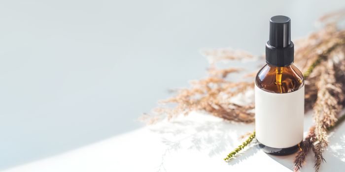 Concept of apothecary cosmetic. Herbs and brown glass bottle of essential oil in still life composition on light blue and white background. Sunlight and shadow. Natural cosmetic remedy with white tag.