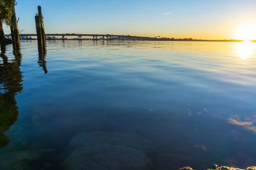 Tauranga Harbour Bridge arches across the harbour in distance back-lit by golden sunrise glow between blue calm water of bay and sky.