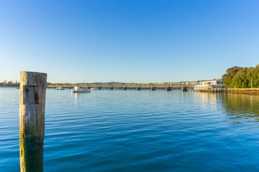 Tauranga Railway bridge catches glow from sunrise in distance across  blue calm water in low level background image.