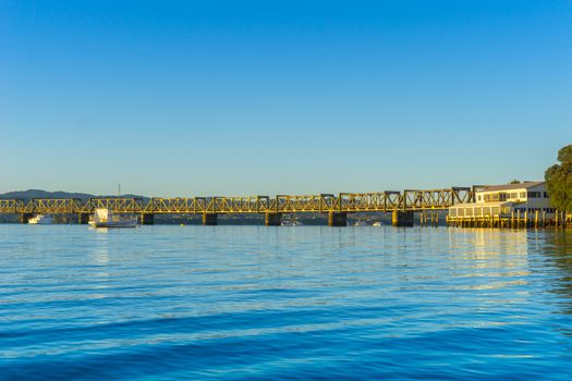 Tauranga Railway bridge catches glow from sunrise in distance across  blue calm water in low level background image.