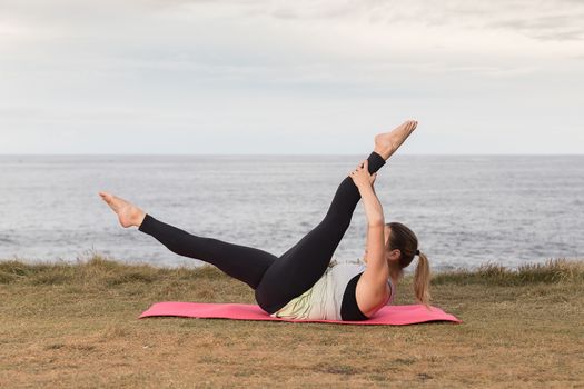 Woman in sportswear doing pilates outdoors on a pink mat with the sea in the background.