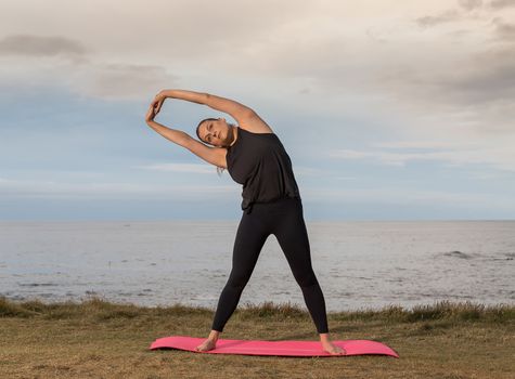 Woman in sportswear stretching outdoors on a pink mat with the sea in the background.