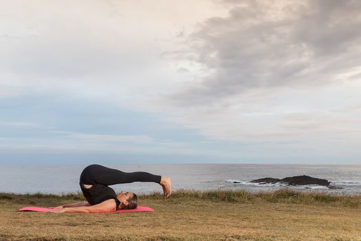 Woman in sportswear doing pilates outdoors on a pink mat with the sea in the background.