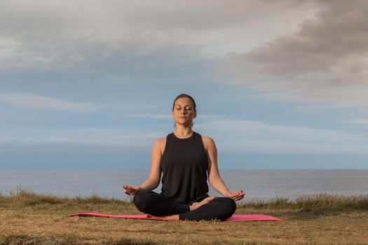 Woman in sportswear doing yoga outdoors on a pink mat with the sea in the background.