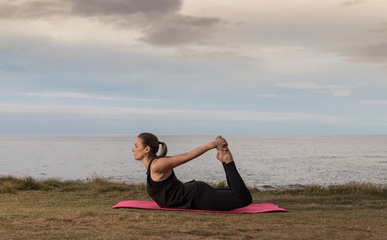 Woman in sportswear doing pilates outdoors on a pink mat with the sea in the background.