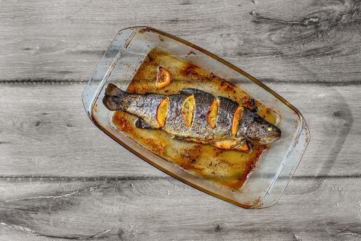 Table top view on grilled trout with orages in glass baking pan, on gray wood table.