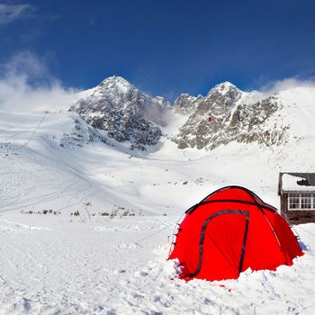 Bright red climbing tent on snow with Lomnicky Stit peak in the background, on a sunny winter day. Tatranska Lomnica ski resort, Slovakia