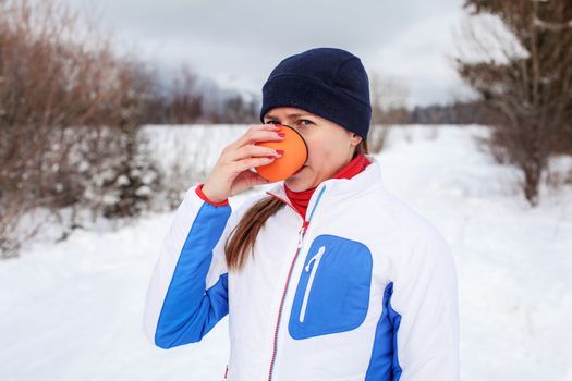 Young woman in winter sport jacket drinking hot tea from orange thermos flask cup on cold overcast day with snow covered landscape in background.