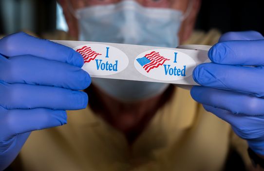 Election volunteer with face mask and medical gloves holding roll of I Voted buttons with USA Flag ready for in person voters in Presidential election