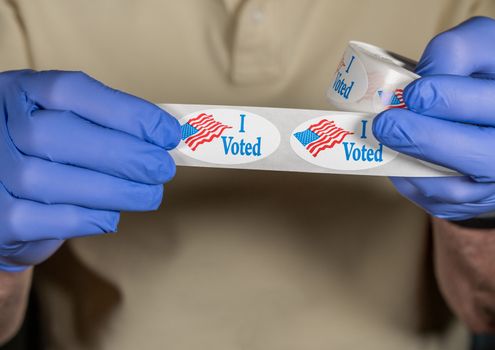 Hands in medical gloves holding roll of I Voted buttons with USA Flag ready for in person voters in Presidential election