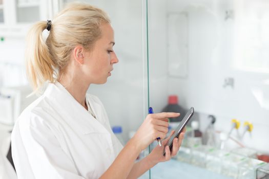 Portrait of young female researcher checking research experiment protocol and making notes on mobile phone application in life science laboratory.