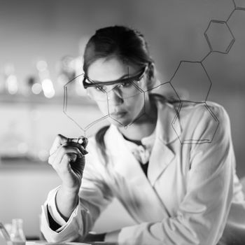 Portrait of a confident female health care expert in life science laboratory writing structural chemical formula on a glass board. Healthcare and modern life science concept. Black and white image.