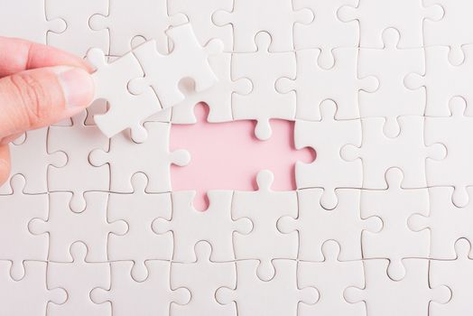 Top view flat lay of hand-holding last piece white paper jigsaw puzzle game last pieces put to place for solve problem complete mission, studio shot on a pink background, quiz calculation concept