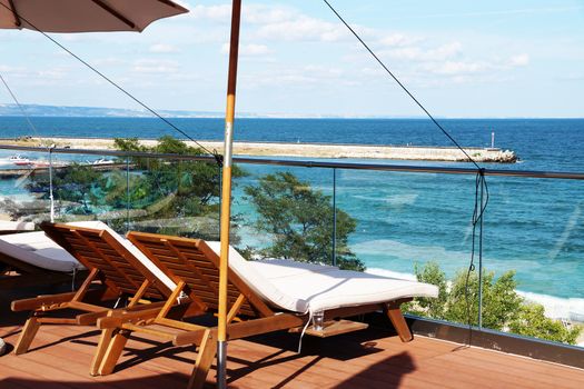 empty sun loungers under an umbrella on the patio against the backdrop of the sea.