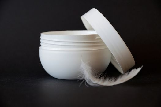 open white jar of cream and white feather on black background, copy space