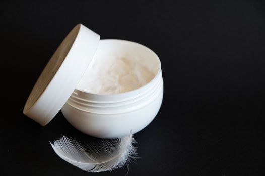 open white jar of cream and white feather on black background, copy space