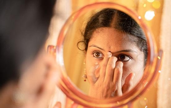 Indian married Woman applying Bindi, sindoor or decorative mark to forehead in front of mirror during festival celebrations with decoration lights as background