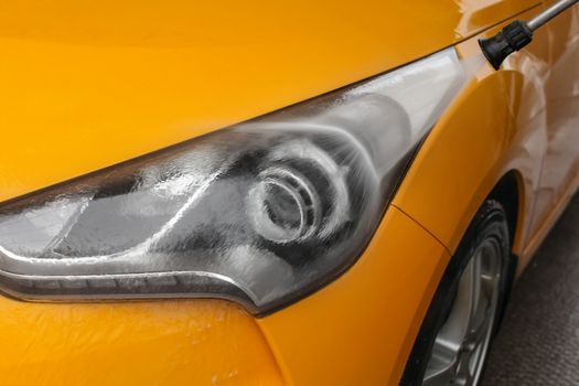 Detail on dark yellow car front light being washed with jet water in carwash.